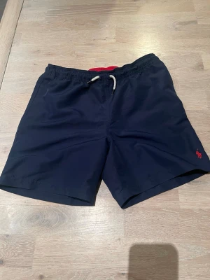 Marinblå badbyxor från Polo Ralph Lauren, size L/14-16y - Snygga marinblå badbyxor från Polo Ralph Lauren med vit dragsko i midjan och röd logga broderad på benet. Insidan har meshfoder och midjan är elastisk för skön passform. Perfekta för stranden eller poolen!