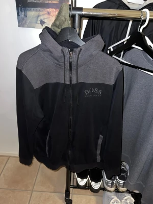 Gamla modellen av Hugo Boss - Snygg hoodie från Hugo boss i svart och grått med dragkedja framtill och huva med logotypdetalj. Mjuk bomullsblandning, två fickor och diskret BOSS-tryck på bröstet. Perfekt för en avslappnad streetstyle-look.