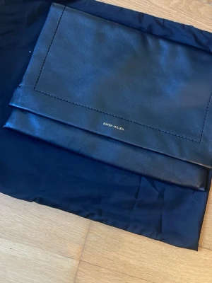 Svart läderhandväska från Karen Millen - Stilren svart handväska i äkta läder från Karen Millen med guldfärgade dragkedjor och diskret rund detalj på framsidan. Väskan har rymligt fack och klassisk form, perfekt för att bära dina essentials. Snygg och tidlös design.