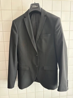 Svart kavaj från Selected Homme - Stilren svart kavaj från Selected Homme med två knappar framtill, klassiska slag och tre fickor. Tillverkad av återvunna fibrer för en mer hållbar stil. Perfekt till skolavslutning/baler mm 