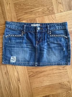 jeanskjol med nitar - Snygg blå jeanskjol med slitningar och nitar vid fickorna. Kjolen är kort, har klassiska fem fickor och bälteshällor. Den är lågmidjad💗