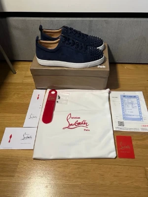 Christian Louboutin sneakers med nitar - Snygga mörkblå sneakers från Christian Louboutin med ikoniska röda sulor och coola nitar framtill. Skorna har snörning och är tillverkade i mocka med vita gummisulor. Perfekta för dig som vill sticka ut med en lyxig och edgy stil.