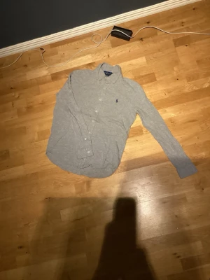 Grå skjorta från Ralph Lauren - Klassisk grå skjorta från Ralph Lauren med knappar framtill och broderad logga på bröstet. Skjortan har lång ärm och normal passform, perfekt till jeans eller chinos. Tillverkad i mjuk bomull för en bekväm känsla. Till ett enkelt billigt pris!  Har även en exakt samma men i mörkblå, skriv vid intresse 