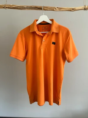 Orange pikétröja från Bogeys & Birdies - Snygg orange pikétröja från Bogeys & Birdies med klassisk krage och knappar framtill. Tröjan har korta ärmar och en liten svart logodetalj på bröstet. Perfekt för en sportig och avslappnad stil.