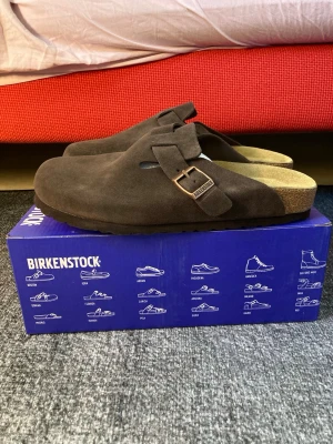 Birkenstock Boston mocka tofflor brun - Säljer ett par klassiska Birkenstock Boston tofflor i mörkbrun mocka. De har en justerbar rem med metallspänne och bekväm fotbädd. Perfekta för dig som gillar stilrena och tidlösa skor.