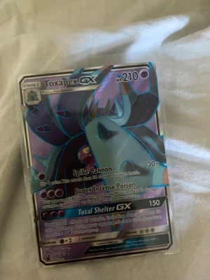 Toxapex GX Pokémonkort - Säljer ett Toxapex GX Pokémonkort med holografisk yta. Kortet har lila och blå färgtoner och är tillverkat i klassiskt Pokémonkort-material. Perfekt för samlare eller dig som vill stärka din lek med en stark GX-kort.