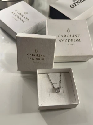 Silverhalsband med klar sten Caroline Svedbom - Ett stilrent halsband i silverfärg från Caroline Svedbom med en gnistrande rund pärla som hänge. Kedjan är tunn och elegant, perfekt för att ge en lyxig touch till din outfit. Levereras i originalask. Säljer då jag bytt från silver till guld.