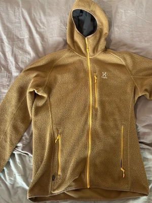 Beige fleecejacka från Haglöfs - Mysig beige fleecejacka från Haglöfs med huva och hel dragkedja framtill. Jackan har två sidofickor med dragkedja och en bröstficka, samt gula detaljer på dragkedjorna. Perfekt för lager-på-lager under kyliga dagar.