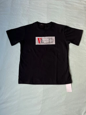 Svart VLONE t-shirt med tryck - Svart t-shirt från VLONE med ett stort rektangulärt tryck på bröstet där det står 'VLONE' i svart och rött mot en grå bakgrund. Klassisk rund hals och korta ärmar. Perfekt för dig som gillar streetwear och vill sticka ut.