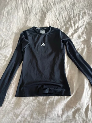 Svart långärmad träningströja Adidas - Säljer en svart långärmad träningströja från Adidas med diskret logga på bröstet. Tröjan är tillverkad i ett stretchigt funktionsmaterial som andas och har en tight passform. Perfekt för löpning, gym eller annan träning.