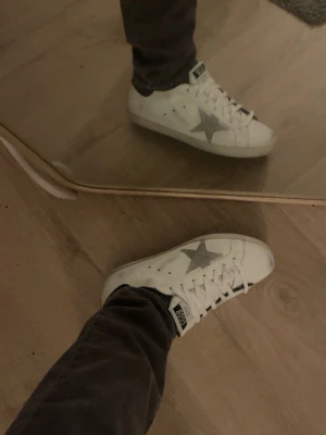 Golden Goose vita sneakers med stjärna - Säljer ett par vita Golden Goose sneakers med grå mockastjärna på sidan och svart detalj vid hälen. Skorna har klassisk låg profil, vita skosnören och perforerade hål för ventilation. Perfekta för en trendig och avslappnad stil.