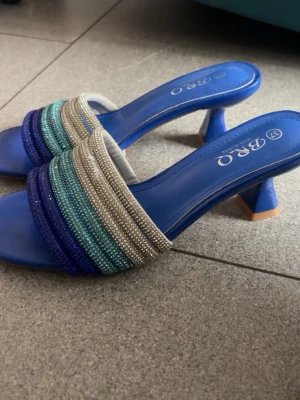 Blåa sandaler med pärldetaljer BRQ - Snygga blåa sandaler från BRQ med låg klack och fyrkantig tå. Ovandelen har flera rader av pärlor i blått, turkost, grönt och silver som ger en glittrig och färgstark look. Perfekta för dig som vill sticka ut med dina skor. Materialet är syntet och pärlor.