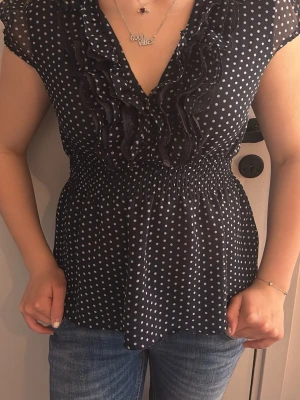 Marin blå prickig blus med volang - Snygg svart blus med vita prickar och volangdetaljer längs den djupa v-ringningen. Blusen har resår i midjan för en smickrande passform och är tillverkad i ett lätt, transparent material. Korta ärmar ger en luftig känsla och toppen passar perfekt till jeans.