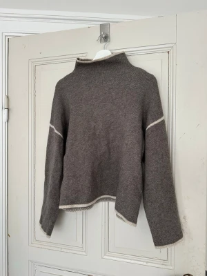 Grå stickad tröja med vita detaljer - Mysig grå stickad tröja med lös passform och hög hals. Tröjan har vita kontrastdetaljer vid ärmslut, nederkant och överarmar. Perfekt för lager-på-lager och chill dagar. Materialet känns mjukt och värmande.