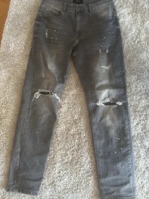 Grå slitna jeans med hål - Säljer ett par grå jeans med skinny fit och slitna detaljer, inklusive stora hål vid knäna och mindre slitningar framtill. Jeansen har klassisk femficksdesign och är perfekta för en avslappnad streetstyle-look.