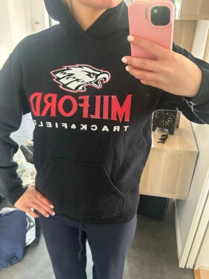 Svart Milford Track & Field hoodie - Säljer en svart hoodie från Milford Track & Field med stor röd och vit logga samt en örn på bröstet. Hoodien har huva och en stor magficka framtill. 