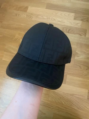 Svart keps  med logomönster - Snygg svart keps  med diskret FF-logomönster över hela kepsen. Klassisk böjd skärm och justerbar rem baktill med gulddetalj. Tillverkad i textilmaterial som ger en lyxig känsla och stilren look.