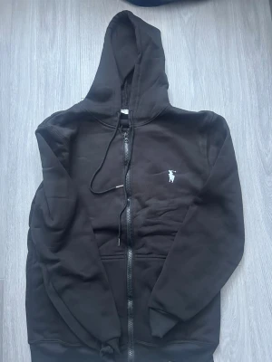 Svart hoodie från Polo Ralph Lauren - Svart hoodie med dragkedja från Polo Ralph Lauren. Klassisk design med vit broderad logga på bröstet, huva med dragsko och två fickor framtill. Tillverkad i mjukt bomullsmaterial som känns skönt mot huden. Perfekt för en avslappnad och stilren look. Storleken är M men skulle även kunna sitta som S. Den är knappt använd därmed har den inga skador eller liknande.