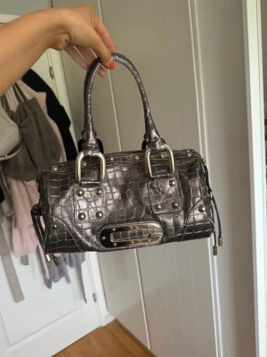 Mörkgrå handväska i krokopräglat läder - Snygg mörkgrå handväska i krokopräglat läder med silverfärgade metalldetaljer och dubbla handtag. Väskan har dekorativa spännen och nitar samt dragkedja upptill. Perfekt accessoar för en trendig look.