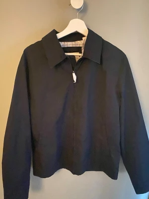 Oanvänd mörkblå minimalistisk vår jacka - Stilren svart jacka med klassisk krage och dold dragkedja framtill. Jackan har raka linjer och enkel design, perfekt för en clean look. Ej använd, lappar kvar. Passar dig som gillar minimalistisk stil.