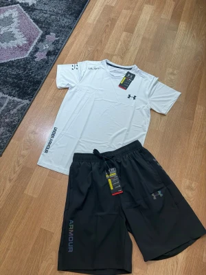 Under Armour träningsset t-shirt & shorts vit/svart storlek M - Snyggt träningsset från Under Armour med en vit t-shirt och svarta shorts. T-shirten har tryckta loggor och textdetaljer, rund hals och kort ärm. Shortsen har elastisk midja med dragsko och logga på benet. Perfekt för gym eller löpning.