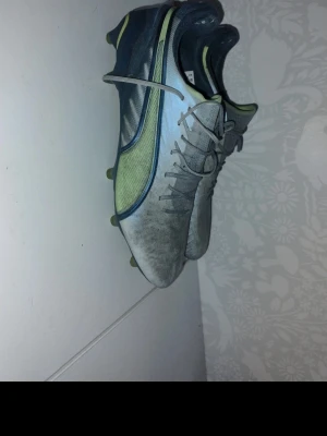 Gråa Puma fotbollsskor med limegrönt - Säljer ett par gråa fotbollsskor från Puma med limegröna och mörkblå detaljer. Skorna har klassisk snörning, låg profil och dobbar för spel på gräs. Ovandelen är i syntetmaterial och designen är sportig och modern.