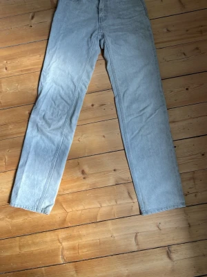 Ljusgrå lågmidbade raka jeans - Säljer ett par ljusgrå jeans med rak passform och klassisk femficksdesign. Jeansen har bälteshällor och stängs med knapp och dragkedja. Perfekta för en avslappnad och stilren look.