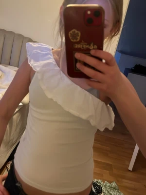 Vit oneshoulder topp med volang - Snygg vit topp med oneshoulder-design och stor volang längs halsringningen. Perfekt för dig som vill sticka ut lite extra. Toppens passform är tight och den är ärmlös på ena sidan. Superfin till både jeans och kjol!