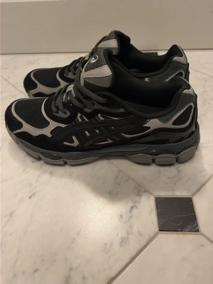 Asics gel Nyc - Snygga Asics sneakers i svart och grått med klassisk mesh och syntetpaneler. Skorna har en chunky sula och ikoniska Asics-stripes på sidorna. Perfekta för dig som gillar sportig stil och vill ha bekväma skor till vardags.