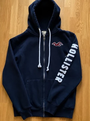 Hollister hoodie - Mörkblå hoodie från Hollister med vit dragkedja och snören i huvan. Stor logga på ena ärmen och klassisk Hollister-fågel på bröstet. Mjuk bomullsblandning och kängurufickor framtill. Perfekt för en avslappnad och stilren look.