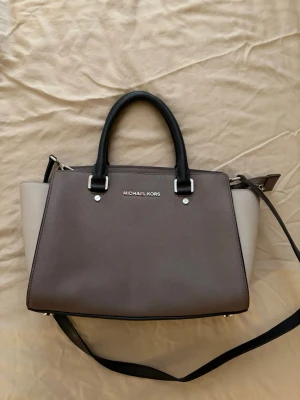 Brun och beige handväska Michael Kors - Snygg handväska från Michael Kors i brunt och beige med svarta handtag och axelrem. Dessvärre ett gammalt märke inuti väskan därav lägre pris, går säkert bort om man försöker 👍🏽