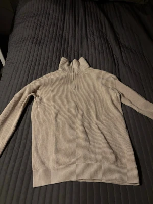 Beige stickad tröja med halv dragkedja - En stilren beige stickad tröja med hög krage och halv dragkedja framtill. Tröjan har långa ärmar och ribbstickade muddar vid ärmslut och nederkant. Perfekt för lager-på-lager och passar till många olika outfits.