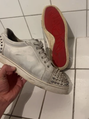 Vita Christian Louboutin sneakers med nitar - Snygga vita sneakers från Christian Louboutin med ikonisk röd sula och coola silverfärgade nitar på tån och hälen. Skorna har klassisk låg modell och är tillverkade i läder med snörning framtill. Perfekta för dig som vill sticka ut med din stil. Tappat X antal spikar men endast en fram resterande bak på