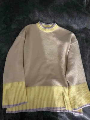 Beige och gul stickad tröja - Säljer en oversized stickad tröja i beige med breda ribbstickade detaljer i ljusgul och lila vid ärmslut, nederkant och halsringning. Tröjan har en avslappnad passform och är perfekt för dig som gillar pastellfärger och mjuka material.