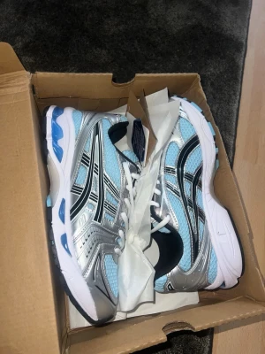 ASICS blå och silver sneakers - Snygga sneakers från ASICS i en fräsch ljusblå färg med silvriga och svarta detaljer. Ovandelen är i mesh och syntetmaterial för bra andningsförmåga och komfort. Sulan är vit med blå inslag och ger en sportig vibe. Perfekta för dig som gillar streetstyle och sportig look.