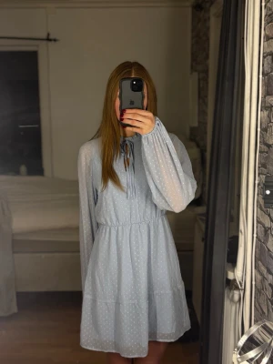 Ljusblå prickig klänning med knyt - Söt ljusblå klänning med vita prickar och knyt i halsen. Klänningen har långärmade, lätt transparenta ärmar och markerad midja. Den är kort i modellen och har en luftig, flowy passform. Perfekt för dig som gillar romantisk och feminin stil.