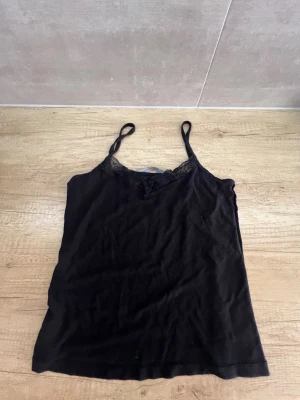 Svart linnetopp med spetsdetalj - Svart linnetopp från Black Label Collection med tunna axelband och snygga spetsdetaljer längs urringningen. Toppen har en liten rosett framtill och är perfekt för en trendig och enkel look.