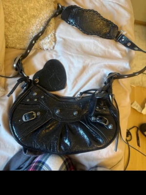 Svart Balenciaga axelväska i skinn - Cool svart handväska från Balenciaga i glansigt skinn med silverfärgade detaljer, dragkedja och spännen. Väskan har en bred axelrem med flätad detalj och en unik form. Perfekt för dig som gillar statement-accessoarer.