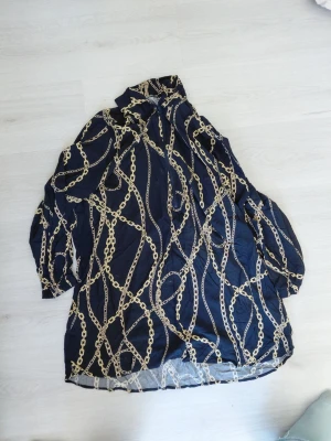 Mönstrad blus från LC Waikiki - Snygg mörkblå blus från LC Waikiki med kedjemönster i guld. Blusen har lång ärm, hög krage och en lös passform. Perfekt för dig som gillar statement-plagg och vill sticka ut med ett coolt print.