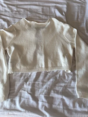 Vit cropped stickad tröja med rosetter - Supersöt vit stickad cropped tröja med tre svarta satängrosetter framtill. Tröjan har långa ärmar och ribbstickade muddar vid ärmslut och nederkant. Perfekt för dig som gillar en gullig och trendig stil.