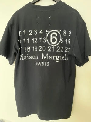 Maison Margiela svart t-shirt - Svart t-shirt från Maison Margiela med ikoniskt tryck av siffror och logga på ryggen. Klassisk passform med korta ärmar och rund hals. Materialet är mjuk bomull som känns skönt mot huden. Perfekt för dig som gillar designerplagg.