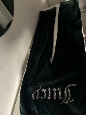 Svarta Juicy Couture mjukisbyxor - Svarta mjukisbyxor i velour från Juicy Couture med glittrande stenar som bildar texten 'Juicy' på ena benet. Byxorna har raka ben och dragsko i midjan för en skön passform. Perfekta för en avslappnad och trendig look. Äkta 