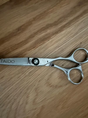 Silvriga frisörsaxar från Taido - Säljer ett par professionella frisörsaxar i silverfärgat rostfritt stål från Taido. Saxarna har ergonomiskt handtag med två fingeröglor och en klassisk, rak design. Perfekta för att klippa och styla håret hemma eller i salong.