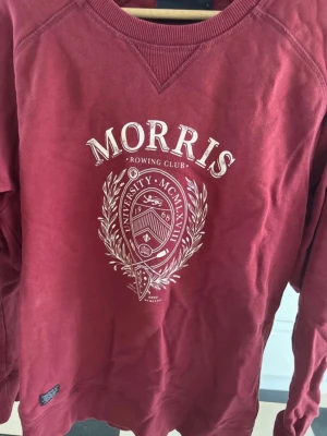 Vinröd sweatshirt från Morris - Snygg vinröd sweatshirt från Morris med tryck framtill som har universitetsinspirerat emblem och texten 'Rowing Club'. Tröjan har rund halsringning, långa ärmar och ribbade muddar. Perfekt för en avslappnad och trendig stil.
