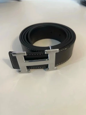 Svart skärp med silver H-spänne - Snyggt svart bälte i skinn med ett stort H-format spänne i silverfärg. Bältet har vita sömmar längs kanterna som ger en stilren kontrast. Perfekt accessoar för att lyfta din outfit.