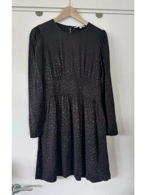 & Other Stories- svart klänning med diskret mönster - storlek 42 - Härlig svart klänning från & Other Stories i storlek 42. Stilren modell med långa ärmar och rund halsringning. Klänningen har puffärmar och en markerad midja med rynkade detaljer. Den har en dold dragkedja i sidan och en knappstängning med fin klädd knapp i nacken. Mönstervävt tyg av små hjärtan i 100% viskos, så bra naturmaterial som andas. Nypris 990:-. Mycket gott skick, som ny!