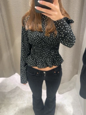 Svart blus med vita prickar - Trendig svart blus med vita prickar och smockad midja som ger en snygg siluett. Blusen har lång ärm och volang nedtill för en extra feminin touch. Perfekt att styla med både jeans och kjol.