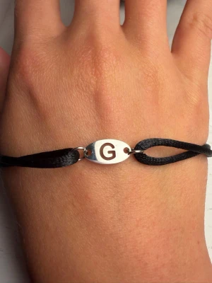Svart armband med bokstav G - Snyggt armband med svart band och en oval silverfärgad bricka i mitten där bokstaven G är ingraverad. Perfekt accessoar för dig som vill ha något personligt och stilrent på handleden. Justerbart band för enkel passform.