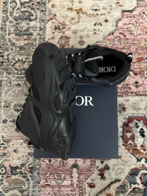 Svarta Dior sneakers med meshdetaljer - Snygga svarta Dior sneakers med chunky sula och meshdetaljer på sidorna. Skorna har en sportig siluett, svarta skosnören och diskret Dior-logga på plösen. Materialmix av läder och mesh ger en modern vibe. Perfekta för dig som vill ha en exklusiv och trendig look.