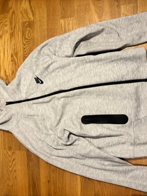 Nike Tech Fleece Zip Hoodie – Grå - Klassisk Nike Tech Fleece i en snygg gråmelerad färg. En tidlös och extremt bekväm hoodie som fungerar lika bra till träning som till en vardagsoutfit. Den har de klassiska Tech Fleece-detaljerna med svarta kontraster och dragkedjefickor. 